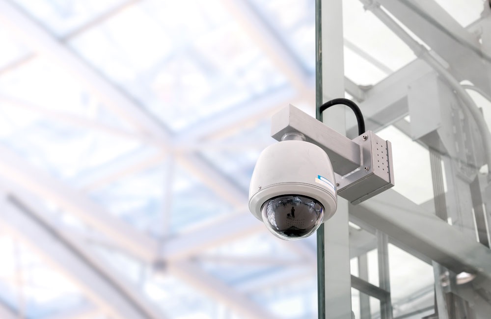 Kopułkowa kamera monitoringu CCTV zamontowana na szklanej ścianie w nowoczesnym budynku z metalową konstrukcją dachu w tle. Widoczny kabel zasilający.