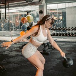 Hornest.gym - Kobieta w sportowym stroju ćwiczy z kettlebell w siłowni w Rybniku. W tle widać sprzęt do ćwiczeń i inne osoby trenujące. Dynamiczne ujęcie w trakcie wykonywania ćwiczenia.