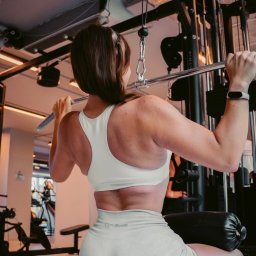 Hornest.gym - Kobieta ćwiczy na maszynie w siłowni, wykonując ćwiczenie na plecy. Widoczny sprzęt Matrix, sportowy strój GymBeam i zegarek na ręce. Ujęcie od tyłu, w jasnych barwach.