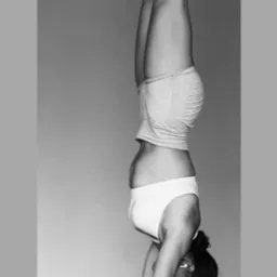 Czarno-białe zdjęcie kobiety w pozycji stania na rękach w minimalistycznym wnętrzu. Widoczne detale: biały top, szorty, skarpety i bransoletka. Upięte włosy.