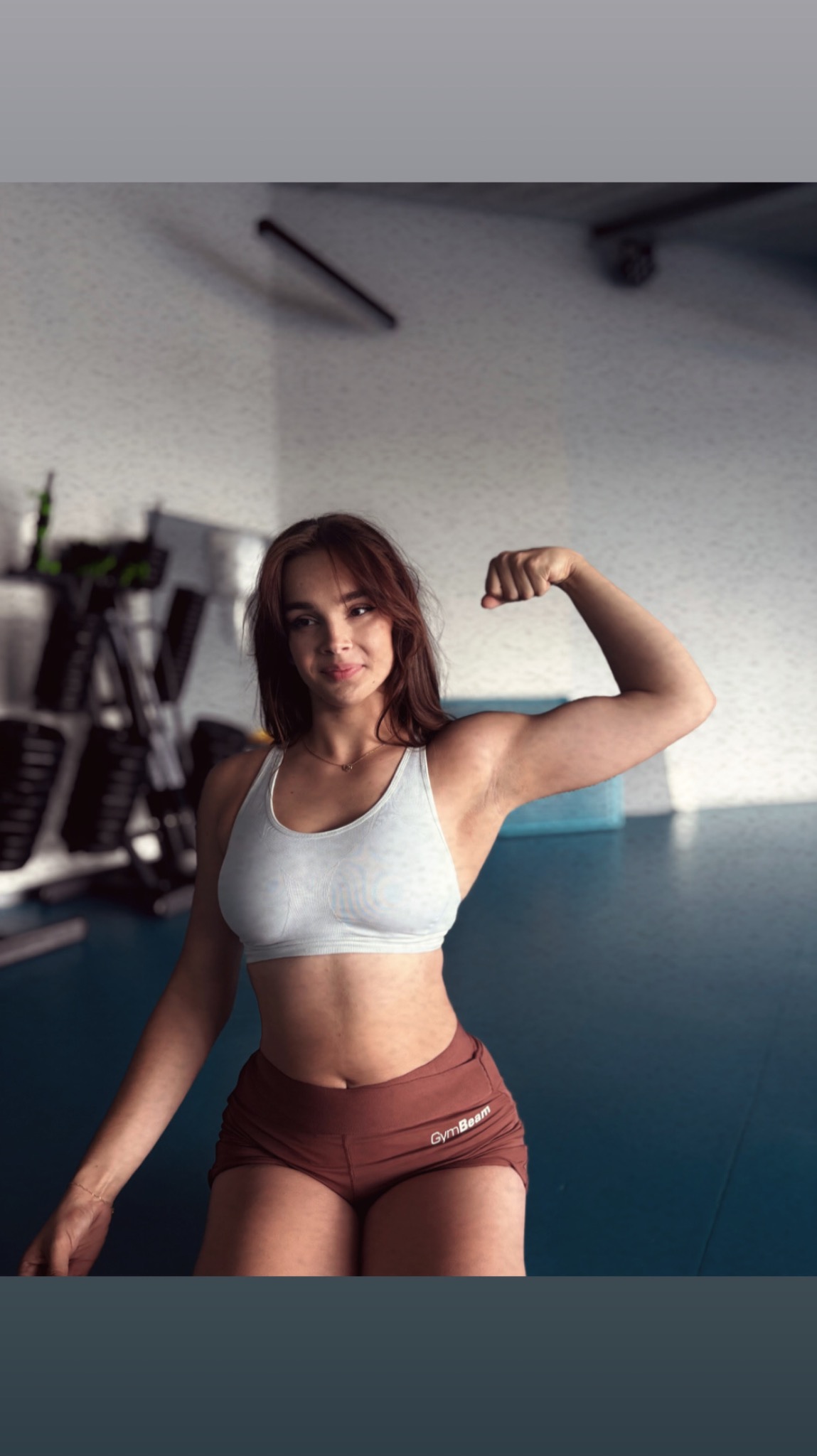 Kobieta w sportowym stroju, prezentująca napięty biceps w siłowni. W tle sprzęt do ćwiczeń i jasne ściany. Dobre oświetlenie podkreśla muskulaturę.