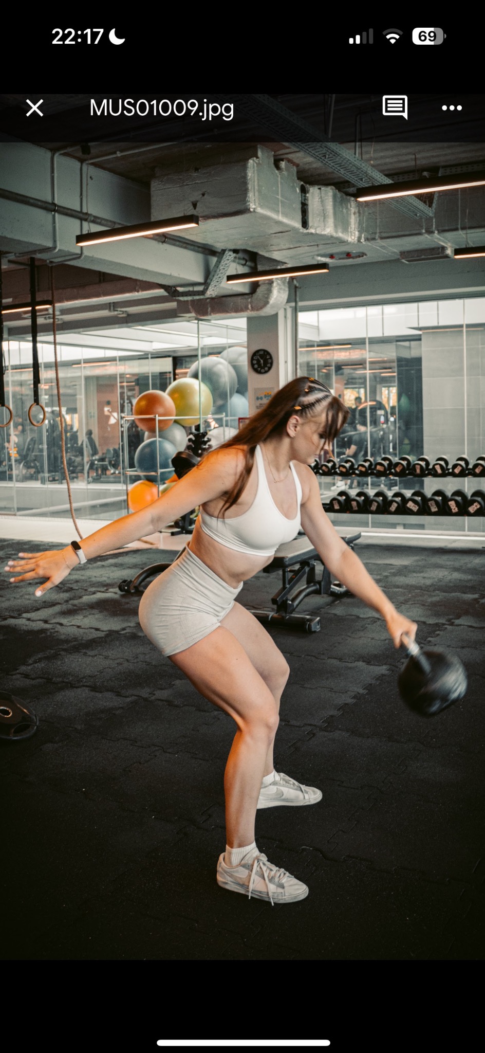 Kobieta w sportowym stroju ćwiczy z kettlebell w siłowni w Rybniku. W tle widać sprzęt do ćwiczeń i inne osoby trenujące. Dynamiczne ujęcie w trakcie wykonywania ćwiczenia.