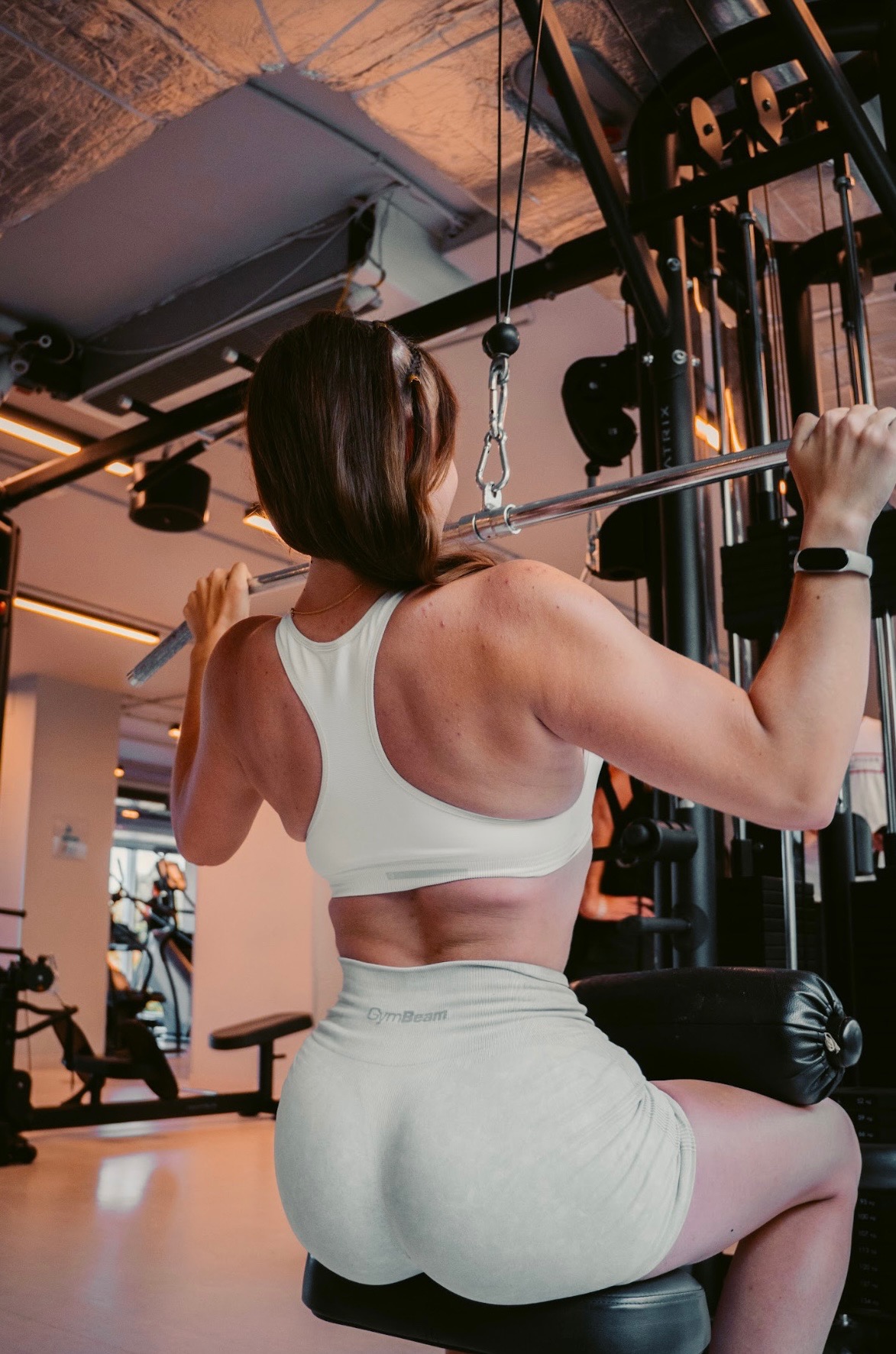 Kobieta ćwiczy na maszynie w siłowni, wykonując ćwiczenie na plecy. Widoczny sprzęt Matrix, sportowy strój GymBeam i zegarek na ręce. Ujęcie od tyłu, w jasnych barwach.