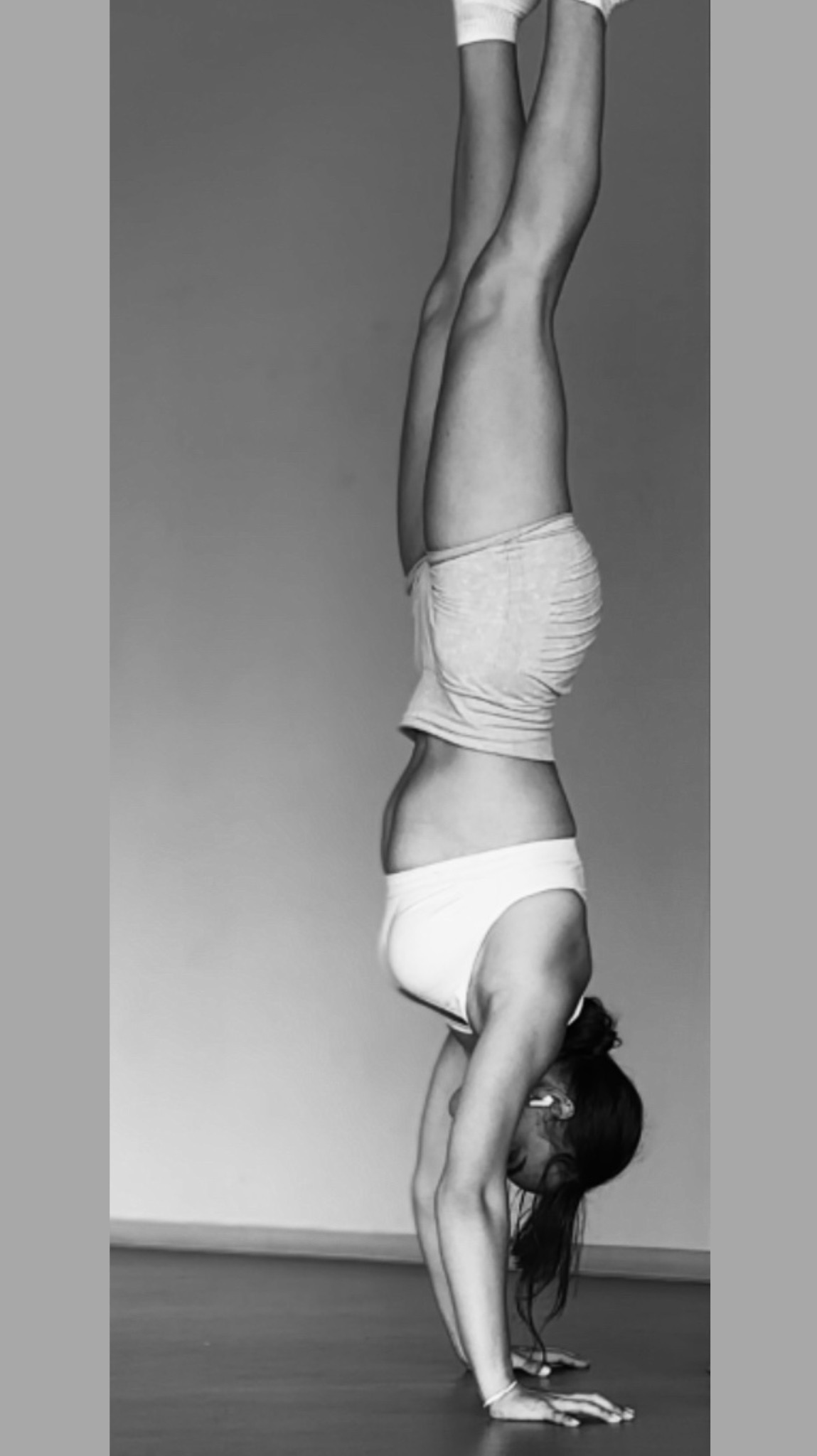 Czarno-białe zdjęcie kobiety w pozycji stania na rękach w minimalistycznym wnętrzu. Widoczne detale: biały top, szorty, skarpety i bransoletka. Upięte włosy.