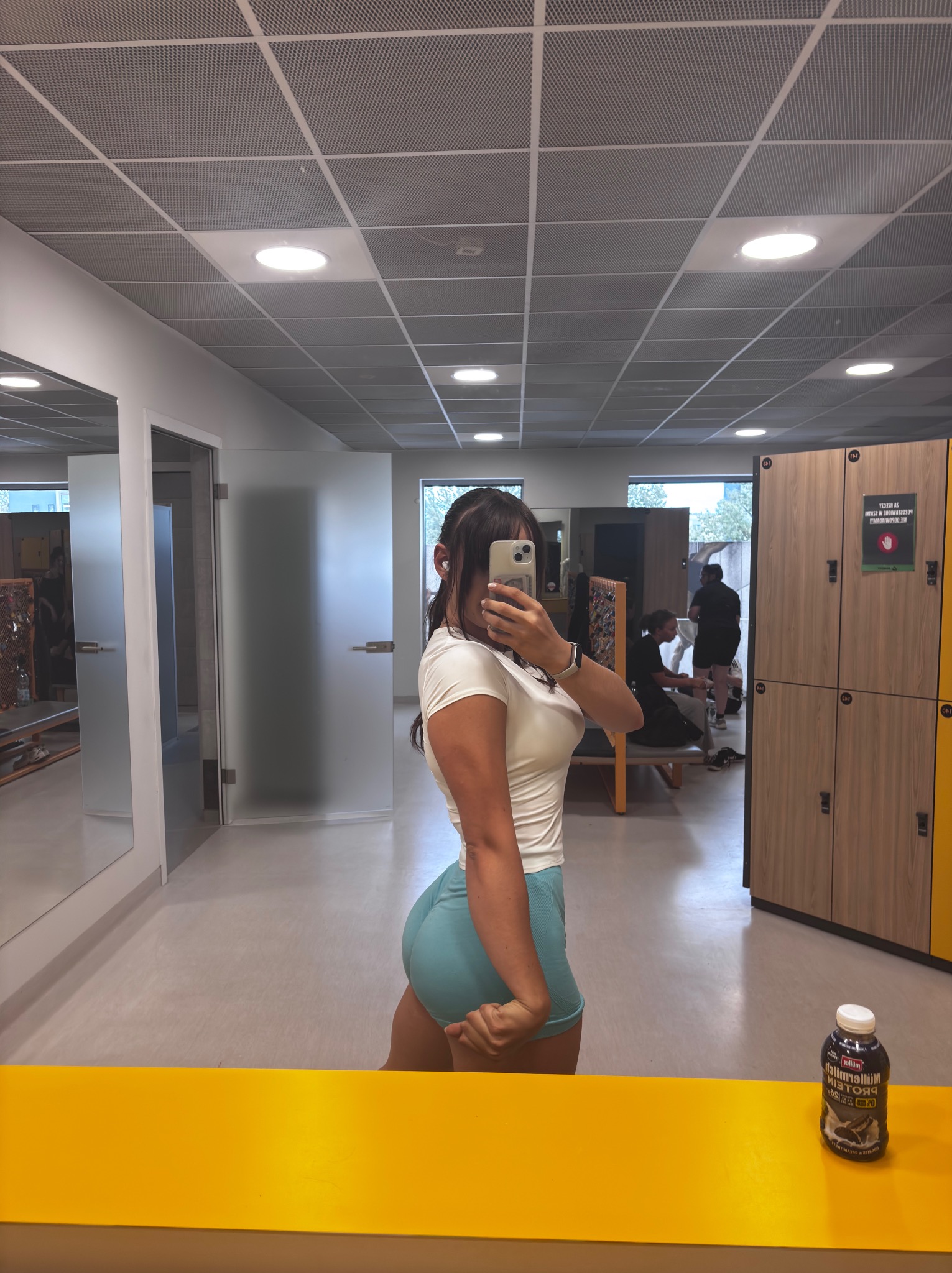 Osoba w sportowym stroju, robiąca sobie selfie w siłowni. Widoczne szafki, lustra i butelka proteinowa. Ujęcie od boku, na pierwszym planie żółty element wyposażenia.