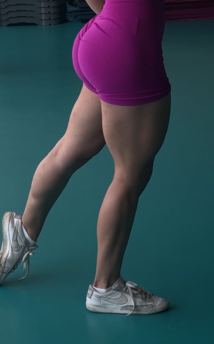 Nogi kobiety w różowych spodenkach i białych butach sportowych, napięte mięśnie nóg, widok z boku na tle turkusowej podłogi, prezentacja efektów treningu.