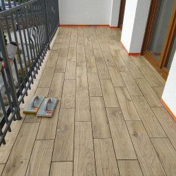 KS Perfect profesjonalne wykończenia wnętrz - Wykończony balkon z drewnopodobną terakotą, zabezpieczony taśmą malarską przy ścianach i oknach. Dwie packi do fugowania leżą na świeżo ułożonej podłodze.