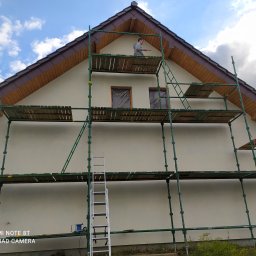 KS Perfect profesjonalne wykończenia wnętrz - Mężczyzna na rusztowaniu maluje elewację domu jednorodzinnego. Widoczne okna zabezpieczone folią, drewniane elementy dachu i drabina.