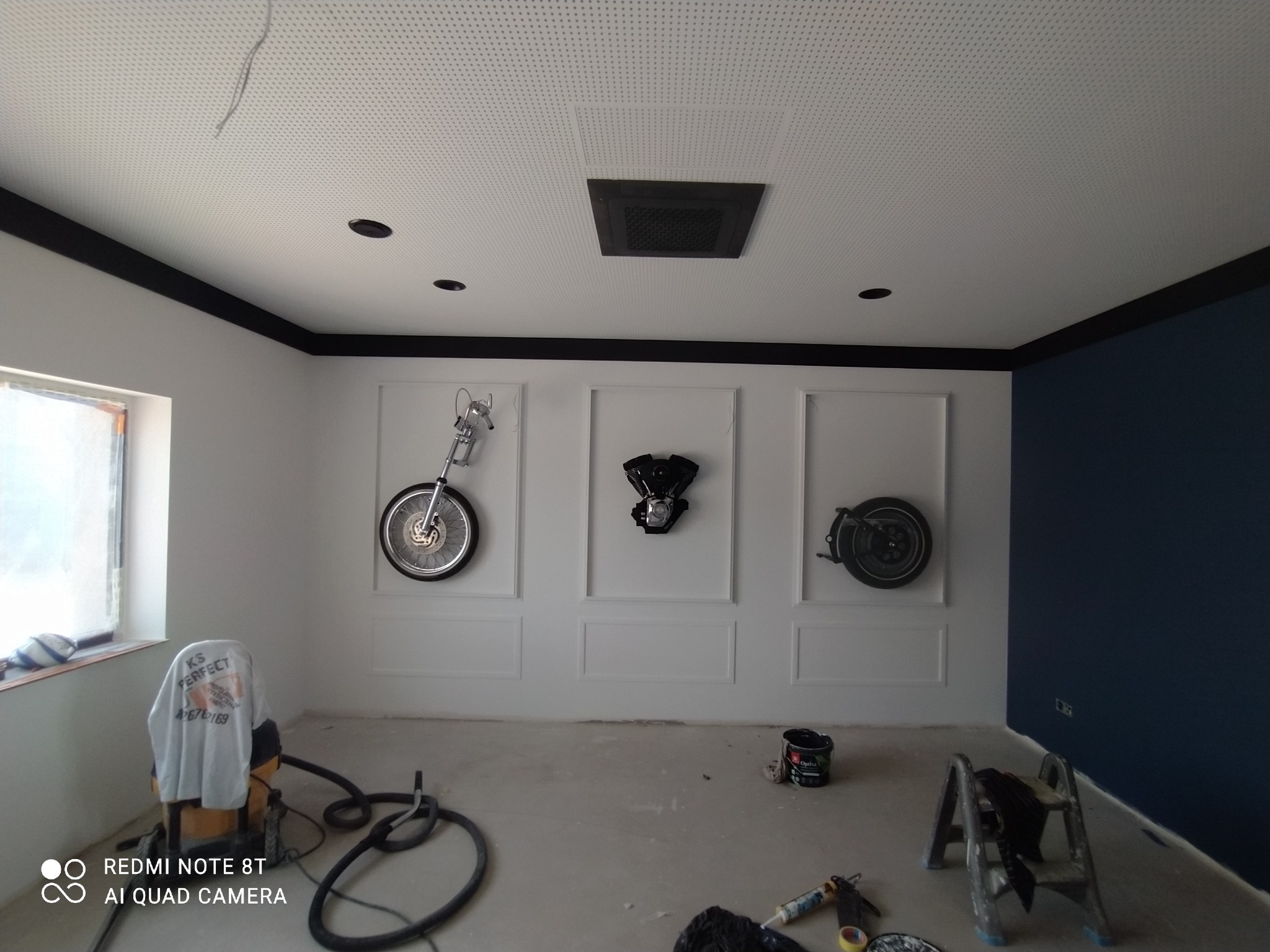 Biało-granatowe wnętrze po remoncie z dekoracjami motoryzacyjnymi na ścianach, widoczny sprzęt malarski i drabina, sufit kasetonowy z czarnymi akcentami.