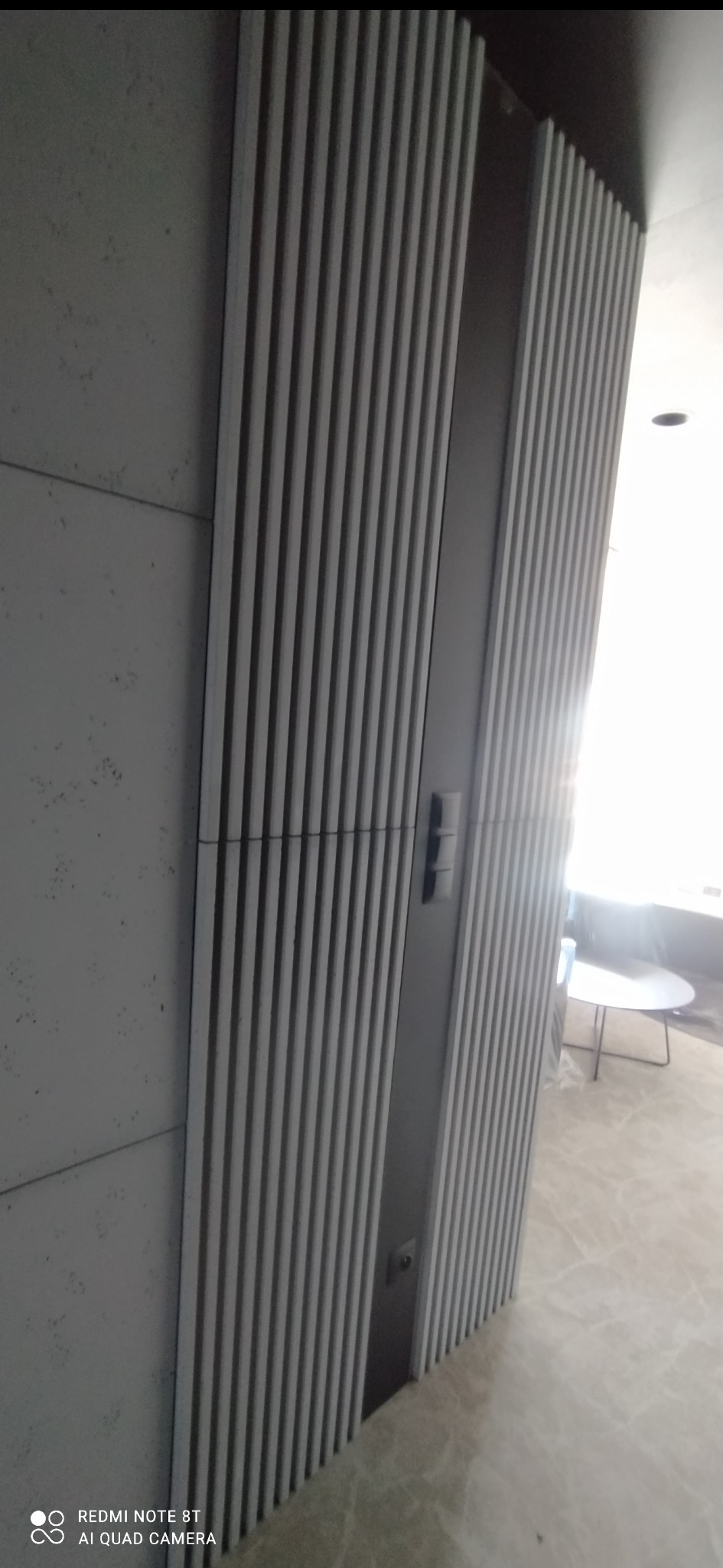 Nowoczesne drzwi z dekoracyjnymi, pionowymi lamelami w odcieniach szarości, z widocznym fragmentem podłogi z marmurowym wzorem. Minimalistyczny design.