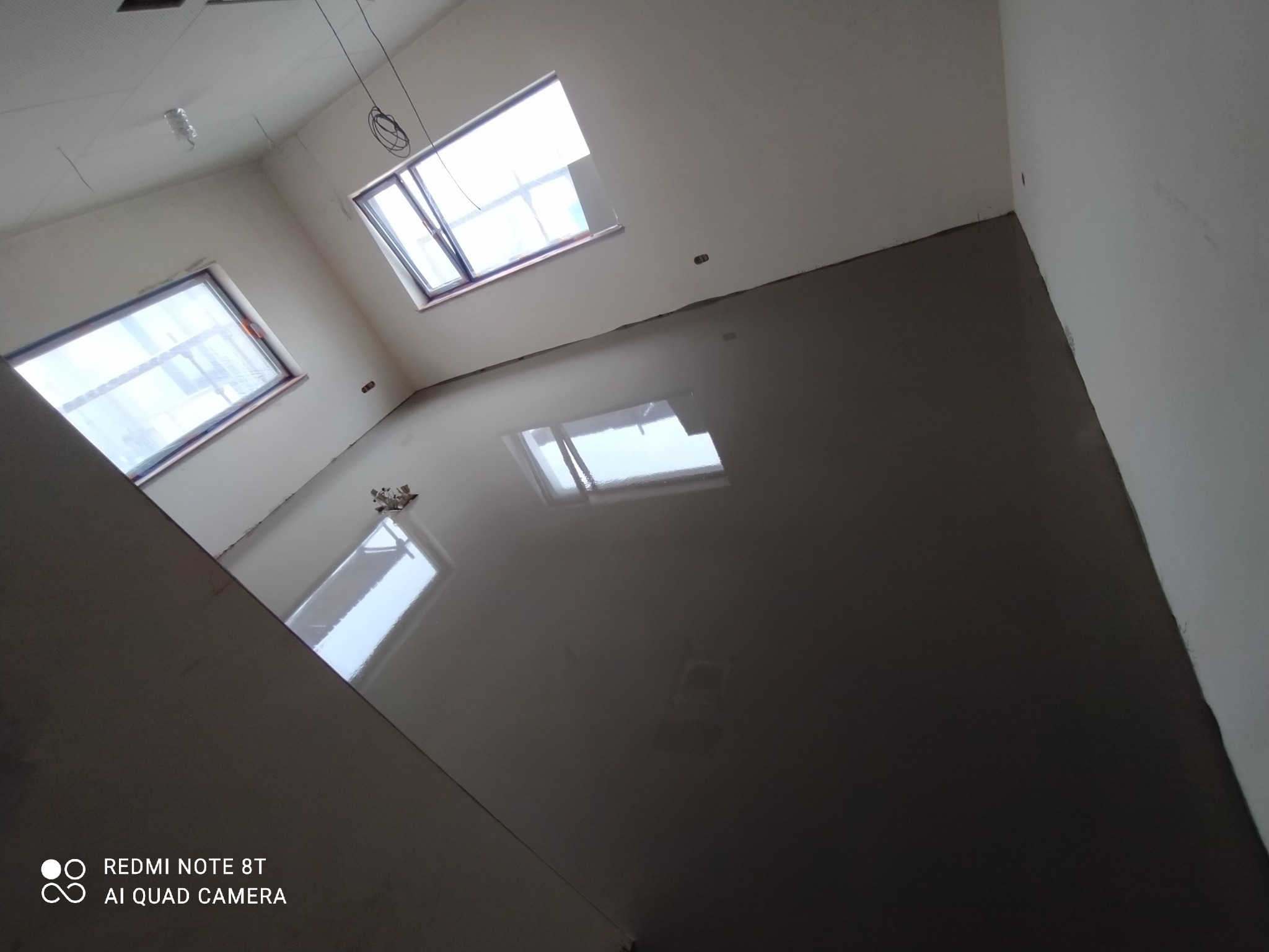 Wnętrze w trakcie wylewki samopoziomującej, widoczne okna i surowe ściany. Nowa, gładka podłoga odbija światło, tworząc lustrzany efekt. Prace wykończeniowe w toku.