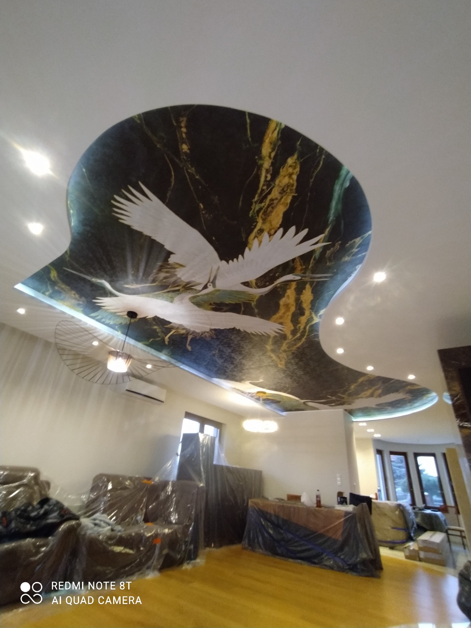 Sufit w salonie z dekoracyjną, marmurkową aplikacją z motywem żurawi w locie. Meble zabezpieczone folią ochronną podczas prac wykończeniowych.