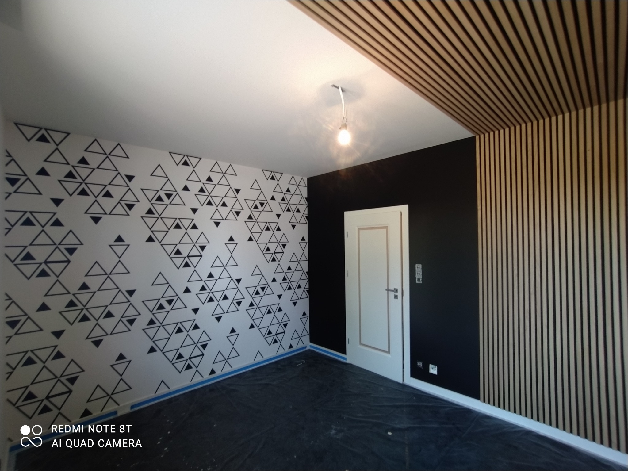 Wnętrze pokoju po remoncie: ściana z deseniem trójkątów, czarna ściana, drewniane lamele na suficie i ścianie, białe drzwi. Oświetlenie punktowe.