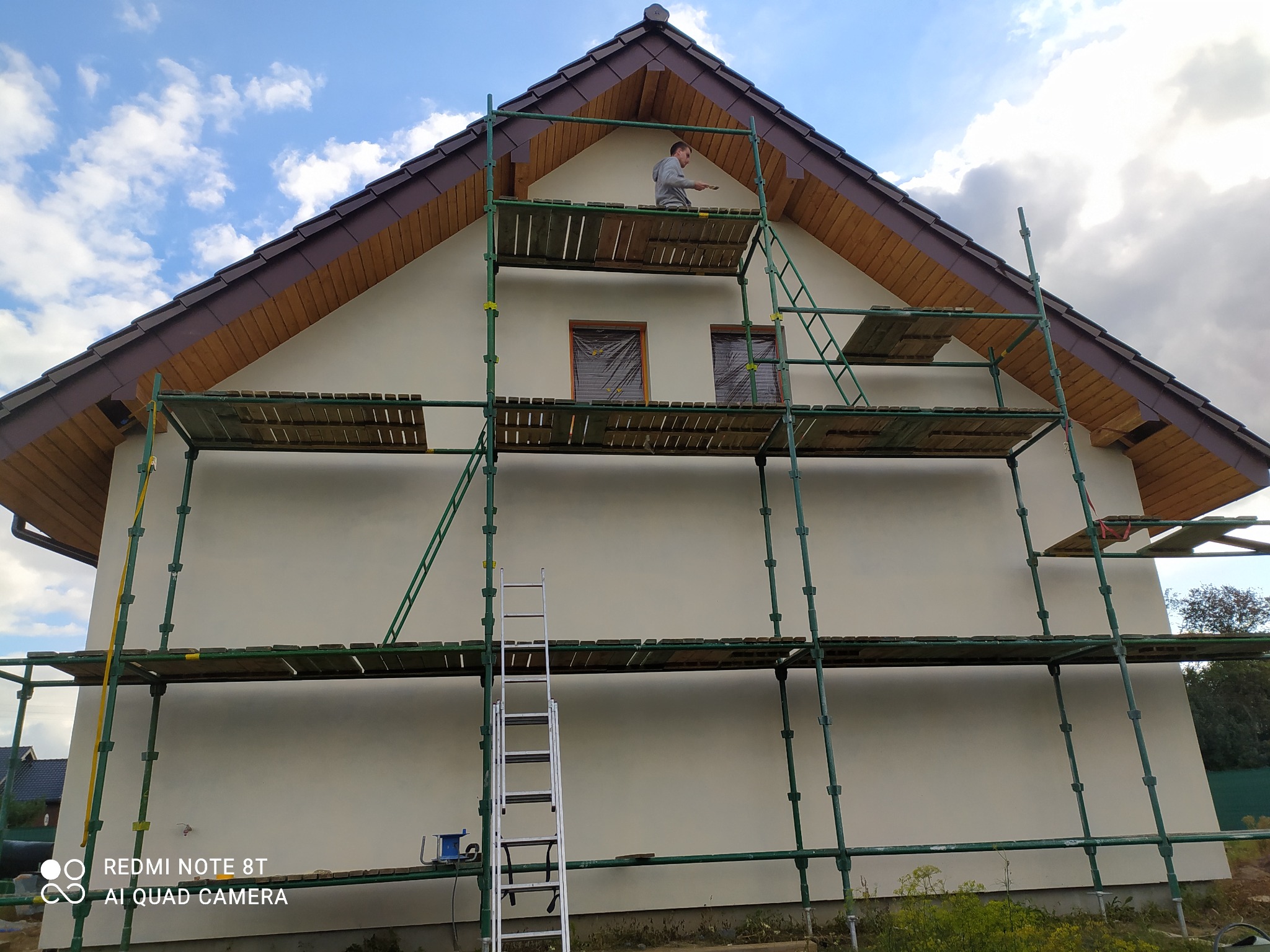 Mężczyzna na rusztowaniu maluje elewację domu jednorodzinnego. Widoczne okna zabezpieczone folią, drewniane elementy dachu i drabina.