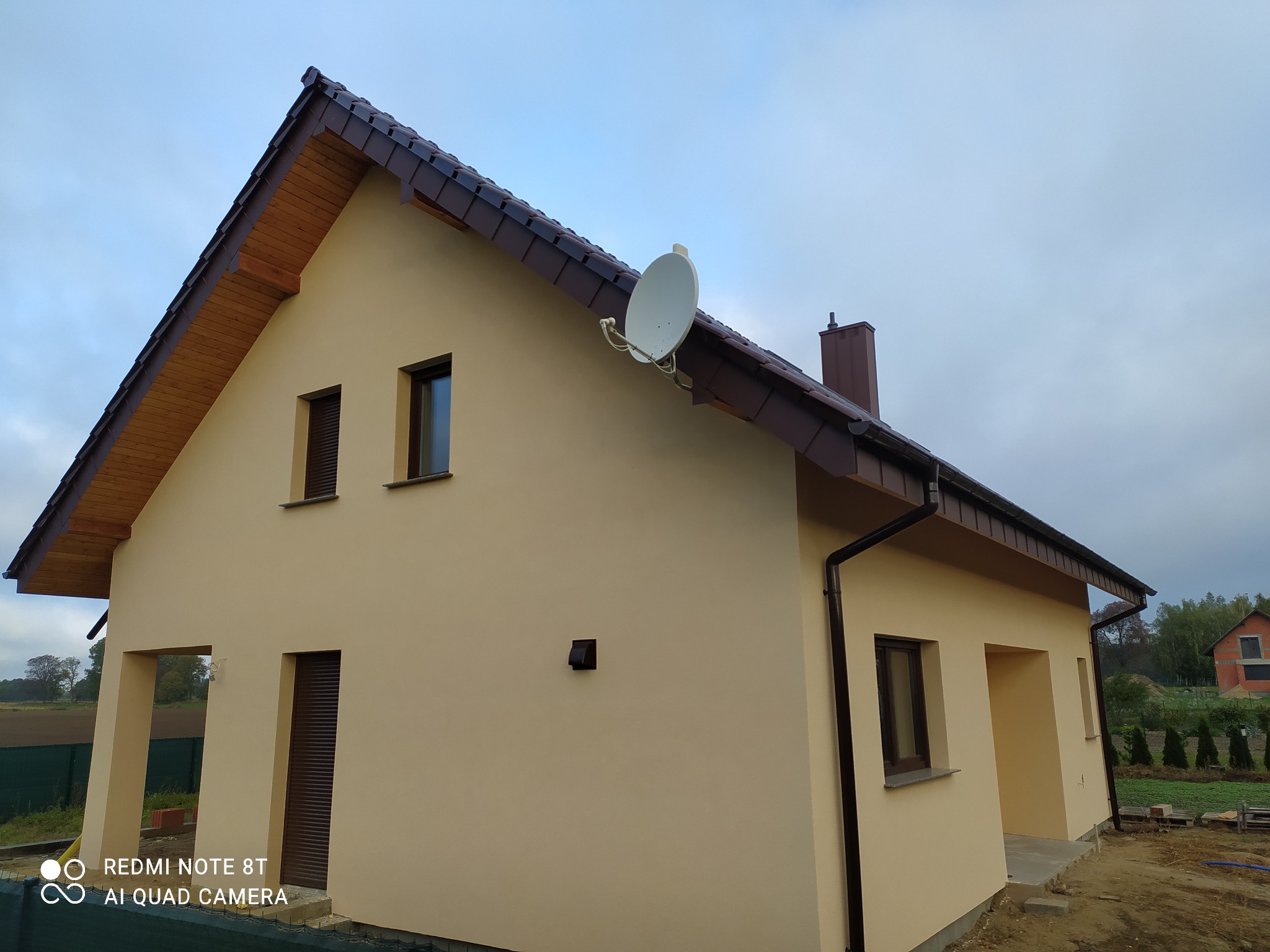 Elewacja domu jednorodzinnego w beżowym kolorze, z brązowymi oknami, dachem i rynnami. Na dachu widoczna antena satelitarna. Dom po remoncie, z nową elewacją.