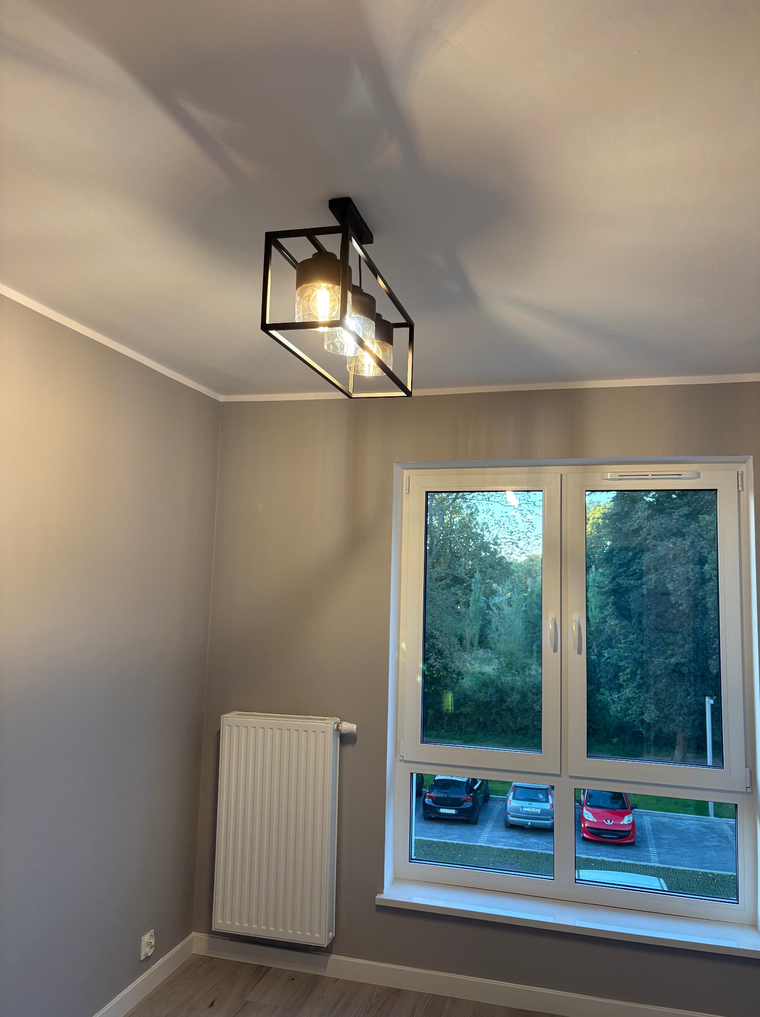 Nowoczesna lampa sufitowa z trzema kloszami w geometrycznej, czarnej oprawie, w jasnym pokoju z oknem na parking. Widoczny grzejnik.