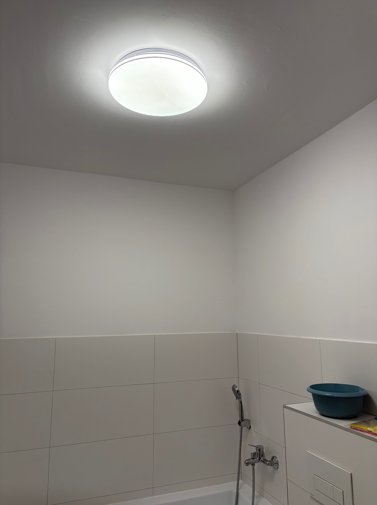 Okrągła lampa sufitowa LED, zamontowana w łazience z białymi płytkami. Widoczny fragment baterii prysznicowej i miska.