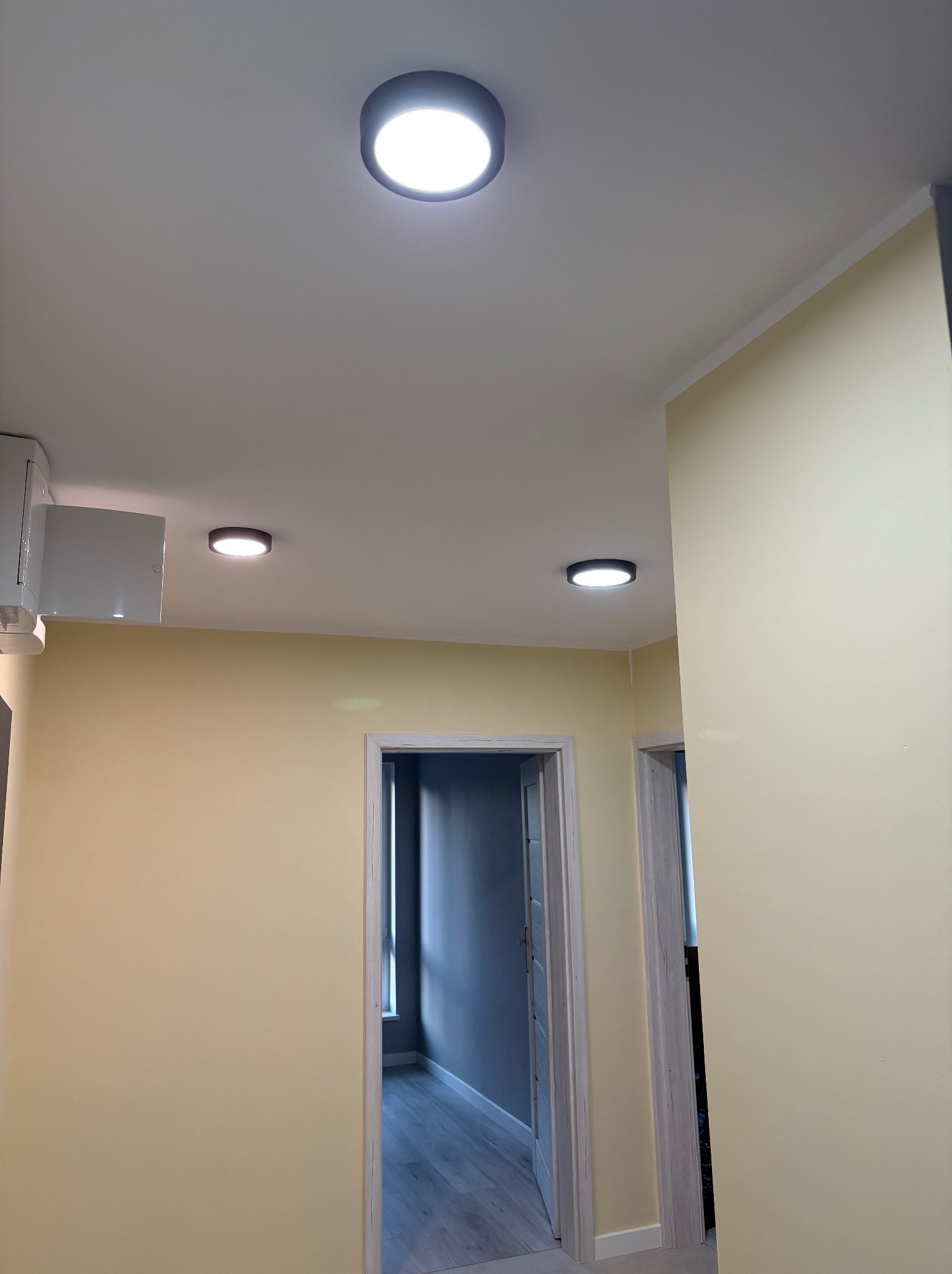 Jasne, okrągłe lampy sufitowe w korytarzu z żółtymi ścianami i dwoma otwartymi przejściami. Minimalistyczny design oświetlenia.