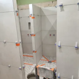 Montaż jasnych płytek ściennych w kabinie prysznicowej, z użyciem klipsów poziomujących. Widoczny fragment podłogi z mozaiką i narzędzia glazurnicze.