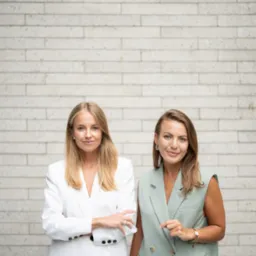 Dwie uśmiechnięte kobiety w eleganckich, dopasowanych kompletach stoją na tle ceglanej ściany. Profesjonalny wizerunek zespołu.