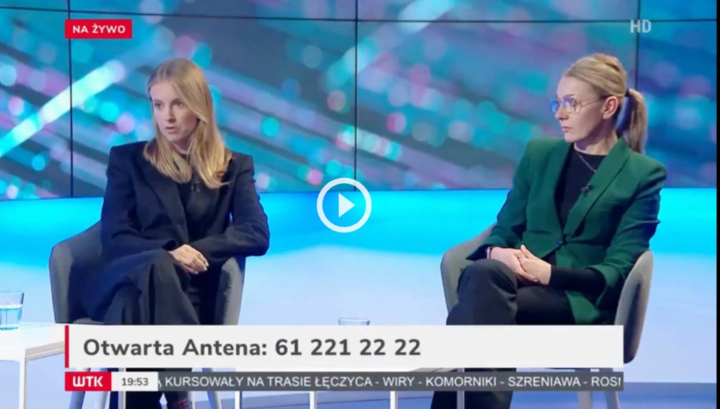 Dwie kobiety w studiu telewizyjnym podczas programu na żywo, siedzące na krzesłach przed ekranem z numerem telefonu. Ubrania w stylu biznesowym.