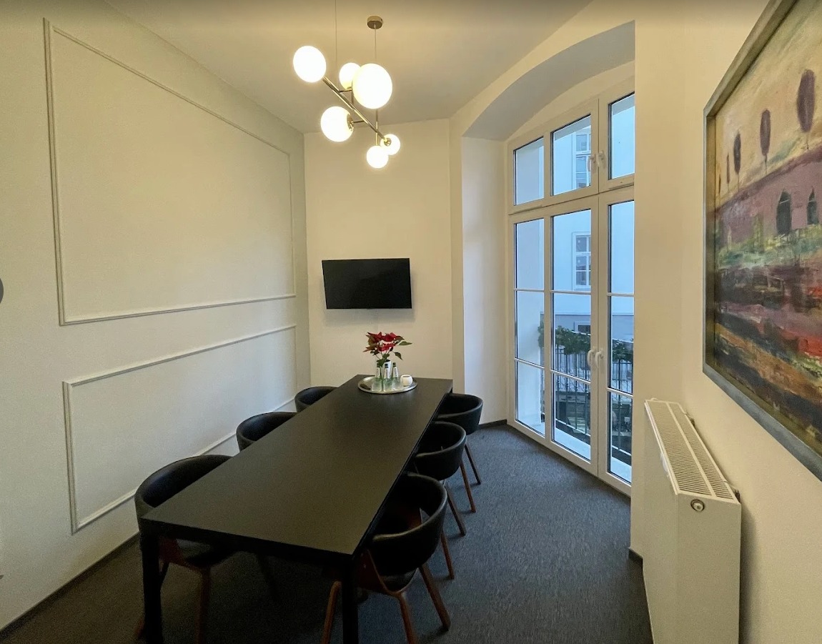 Sala konferencyjna z długim, czarnym stołem i czarnymi krzesłami. Na ścianie telewizor i obraz. Duże okno z widokiem na zewnątrz.