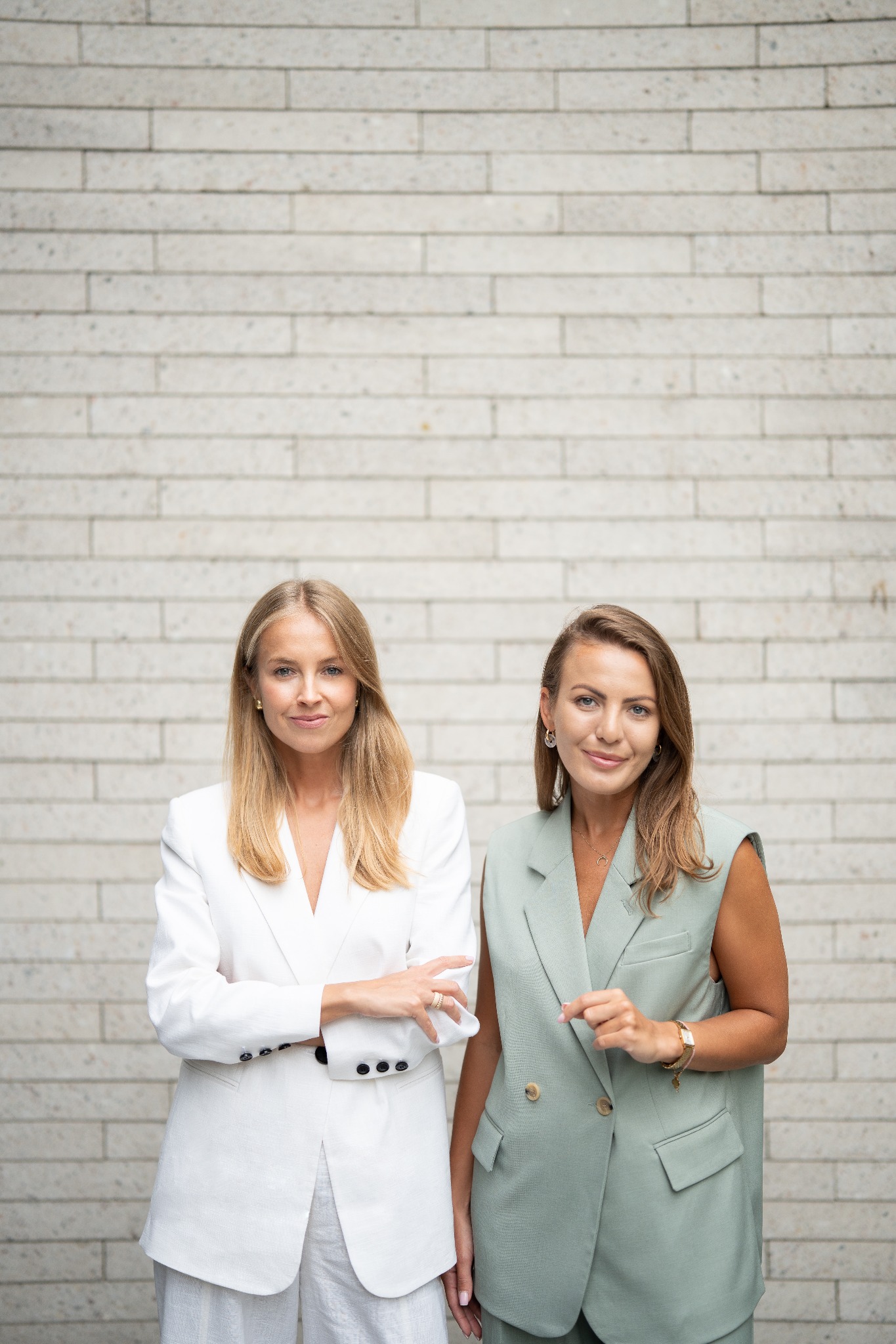 Dwie uśmiechnięte kobiety w eleganckich, dopasowanych kompletach stoją na tle ceglanej ściany. Profesjonalny wizerunek zespołu.