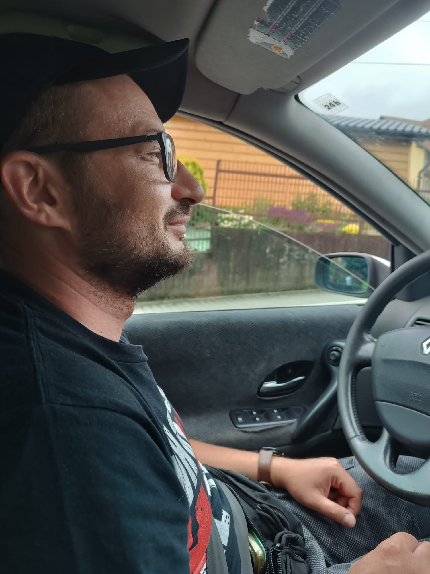 Mężczyzna w okularach i czapce siedzi za kierownicą samochodu, spoglądając w bok. Widoczna deska rozdzielcza i kierownica. W tle zabudowania.