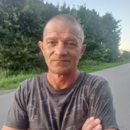 Portret mężczyzny z założonymi rękami na tle drogi i drzew. Widoczne zmarszczki na twarzy i krótko ostrzyżone włosy. Ubrany w szarą koszulkę z wzorem.