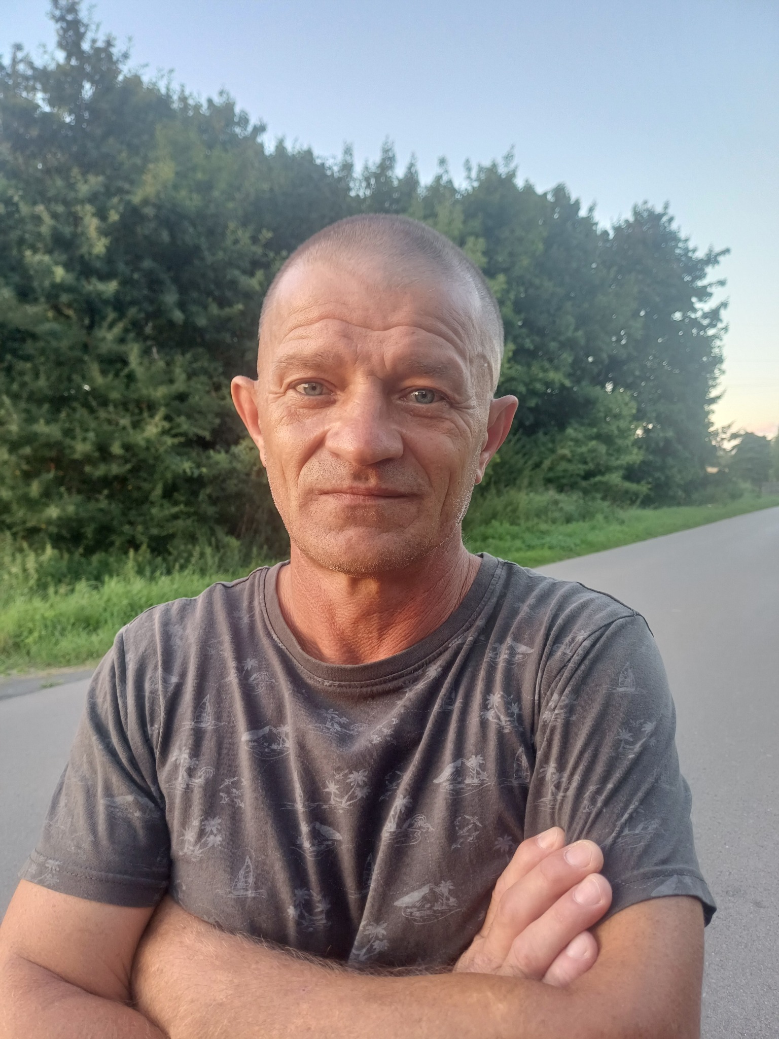 Portret mężczyzny z założonymi rękami na tle drogi i drzew. Widoczne zmarszczki na twarzy i krótko ostrzyżone włosy. Ubrany w szarą koszulkę z wzorem.