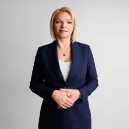 Profesjonalny portret kobiety w granatowym garniturze na jasnym tle. Złoty łańcuszek i pierścionki dodają elegancji. Uśmiech i pewny wyraz twarzy.