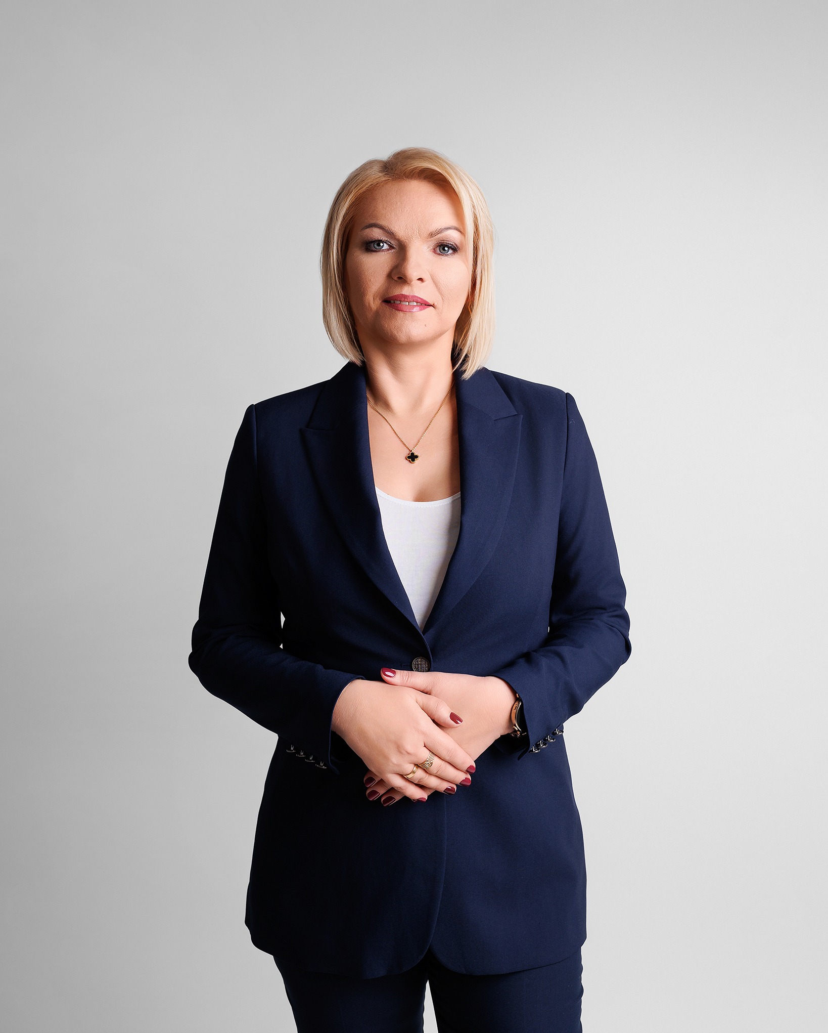 Profesjonalny portret kobiety w granatowym garniturze na jasnym tle. Złoty łańcuszek i pierścionki dodają elegancji. Uśmiech i pewny wyraz twarzy.