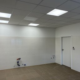 TOMASZ DUSZAK TEAM - Wykończone wnętrze: białe płytki ścienne, brązowa podłoga, sufit podwieszany z oświetleniem. Widoczne wystające rury instalacji wodnej. Drzwi po prawej.