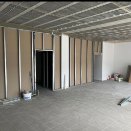 TOMASZ DUSZAK TEAM - Wnętrze w trakcie montażu ścian działowych z płyt gipsowo-kartonowych na metalowym stelażu. Widoczne narzędzia i materiały budowlane. Szara podłoga z płytek.