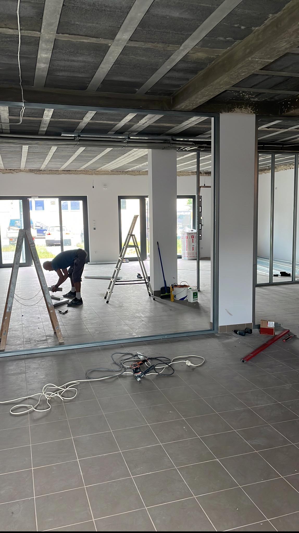 Montaż ścianek działowych z profili stalowych w surowym wnętrzu. Widoczny pracownik przy pracy, drabiny, narzędzia i materiały budowlane. Szare płytki podłogowe.