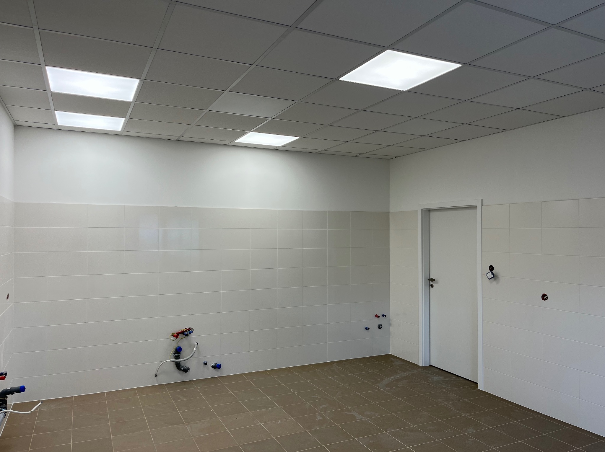 Wykończone wnętrze: białe płytki ścienne, brązowa podłoga, sufit podwieszany z oświetleniem. Widoczne wystające rury instalacji wodnej. Drzwi po prawej.