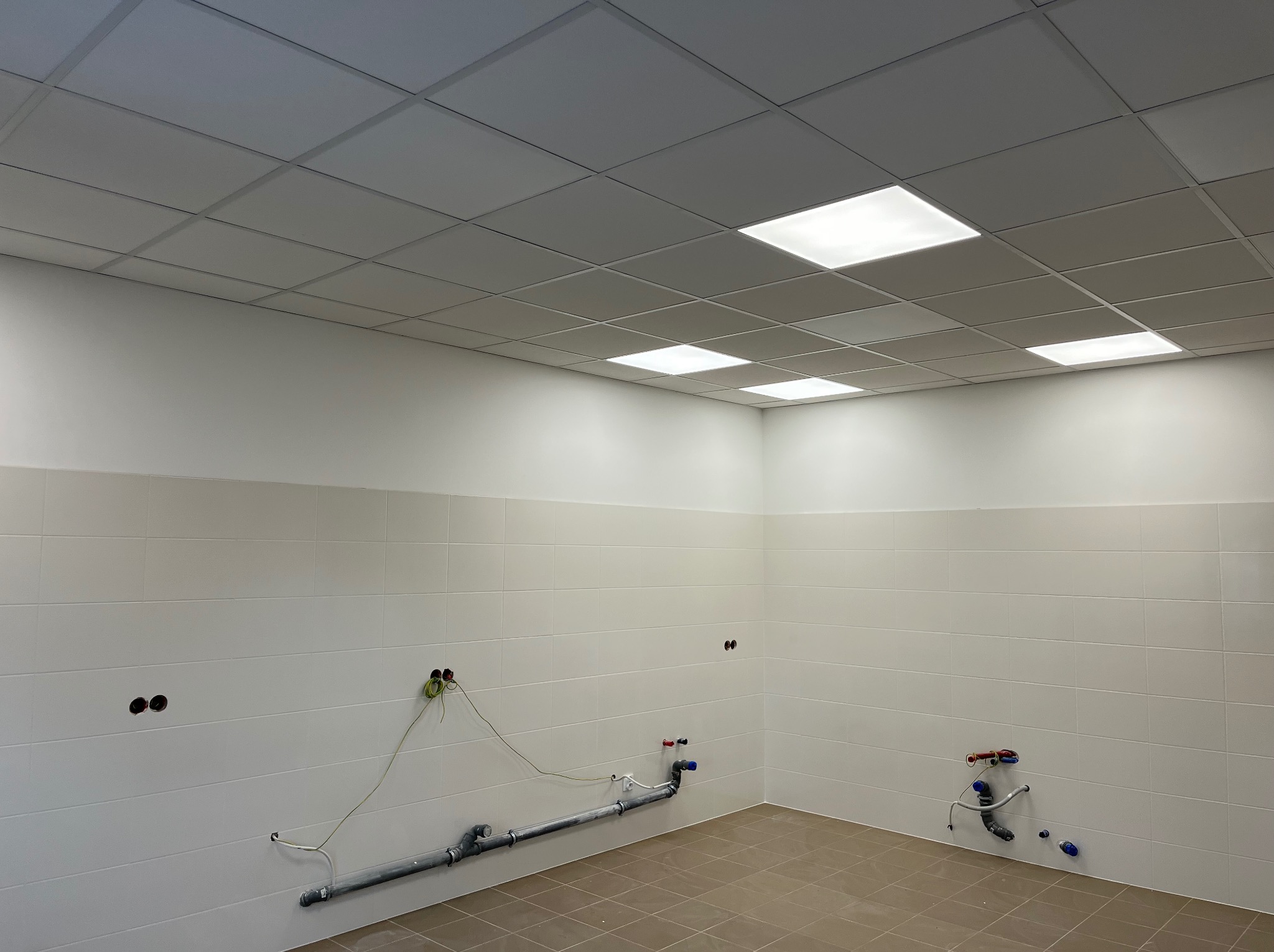 Płytki ścienne i podłogowe w jasnym pomieszczeniu z instalacją hydrauliczną w Poznaniu. Sufit kasetonowy z oświetleniem. Widoczne otwory na gniazdka elektryczne.