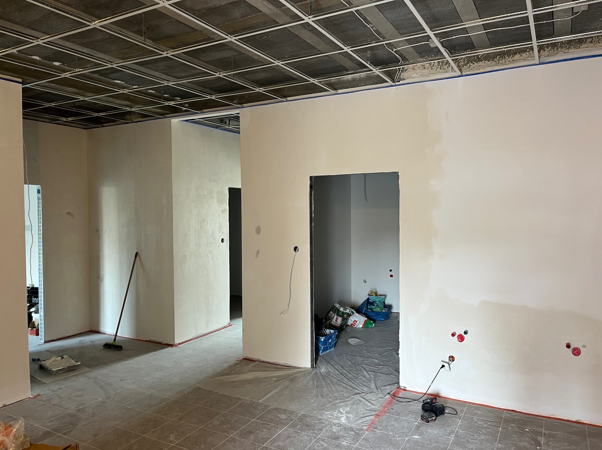 Wnętrze w trakcie prac wykończeniowych: zagruntowane ściany, odkryty sufit z instalacjami, zabezpieczona podłoga i widoczne narzędzia malarskie.