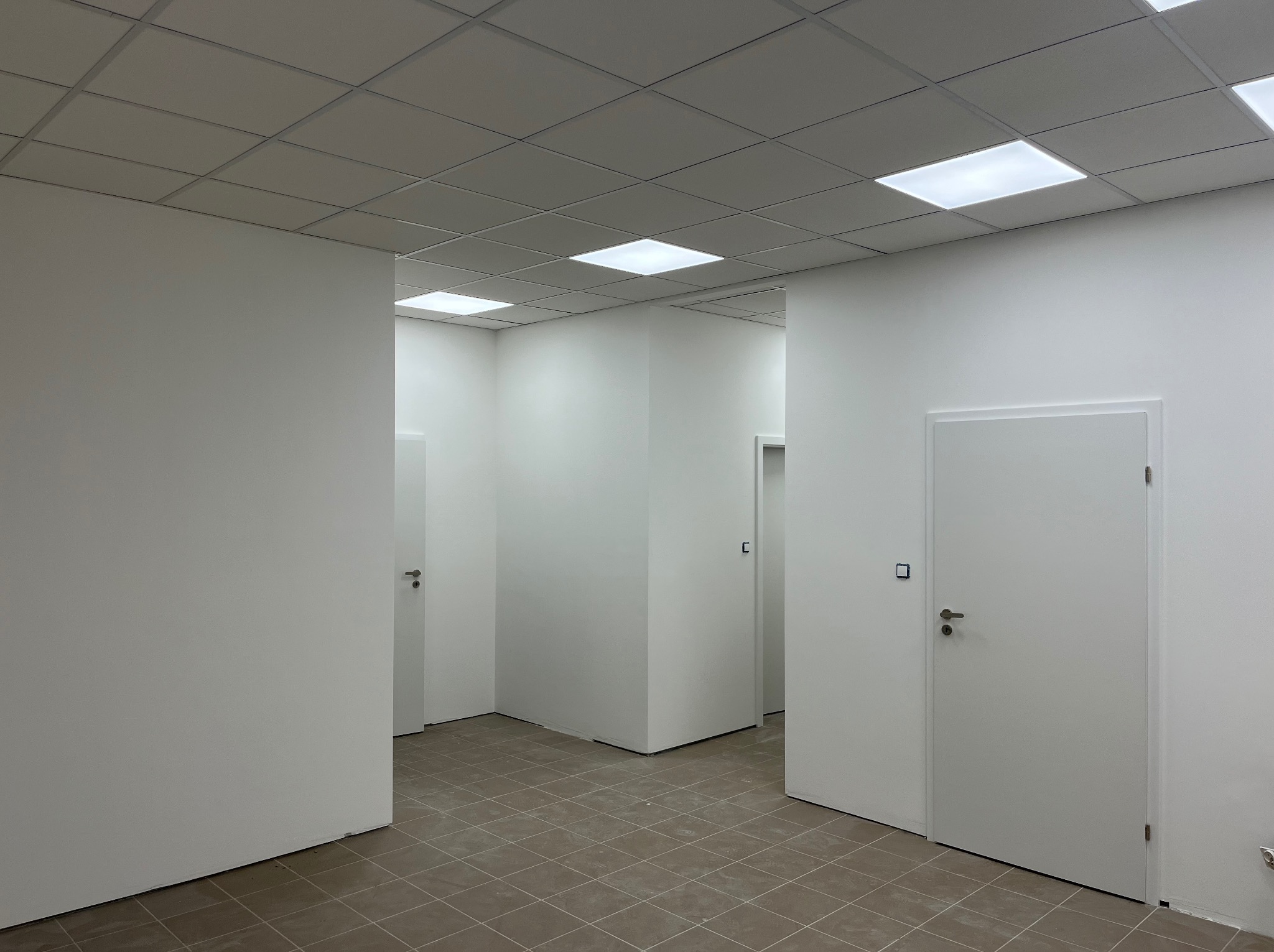 Białe, minimalistyczne wnętrze biurowe z szarą podłogą w kratkę, białymi ścianami, drzwiami i sufitami podwieszanymi z wbudowanym oświetleniem LED.