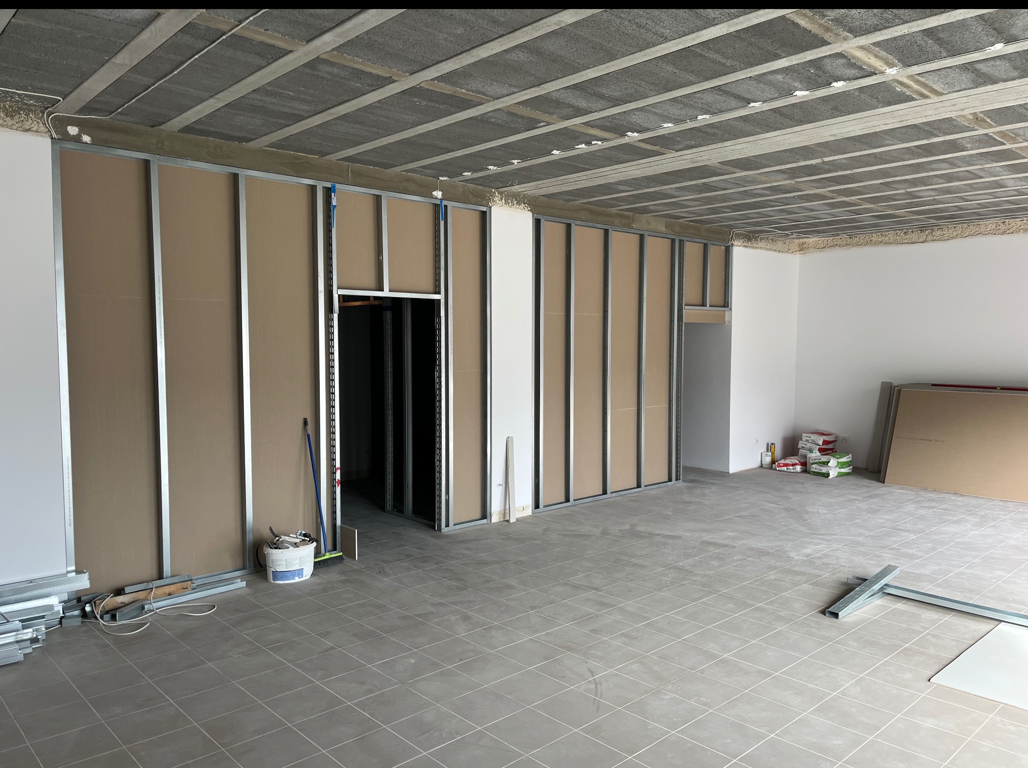 Wnętrze w trakcie montażu ścian działowych z płyt gipsowo-kartonowych na metalowym stelażu. Widoczne narzędzia i materiały budowlane. Szara podłoga z płytek.