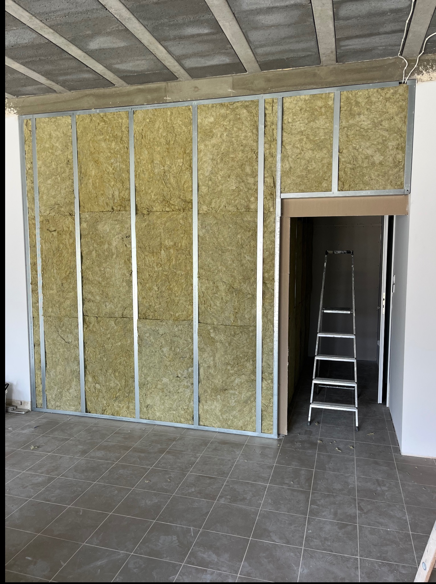 Wnętrze w trakcie wykańczania: ściana z metalowym stelażem wypełniona wełną mineralną, fragment sufitu i podłogi z płytek. W tle drabina w otworze drzwiowym.