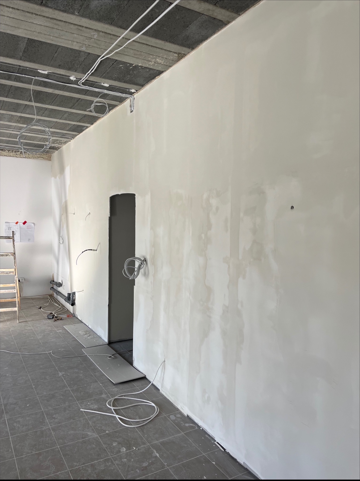 Wnętrze w trakcie gładziowania ścian, widoczne przewody elektryczne i rury. Ściany w kolorze jasnym, sufit z betonu komórkowego. Na podłodze szare płytki.
