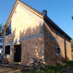 Surowy, dwukondygnacyjny dom z czerwonej cegły, z czarnymi oknami i drzwiami, na tle zieleni i błękitnego nieba. Rower oparty o ścianę. Widoczny etap budowy.