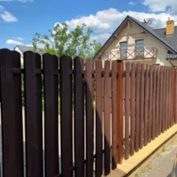 Ciemnobrązowe, drewniane ogrodzenie sztachetowe z jasną podmurówką na tle domu jednorodzinnego i błękitnego nieba z chmurami.