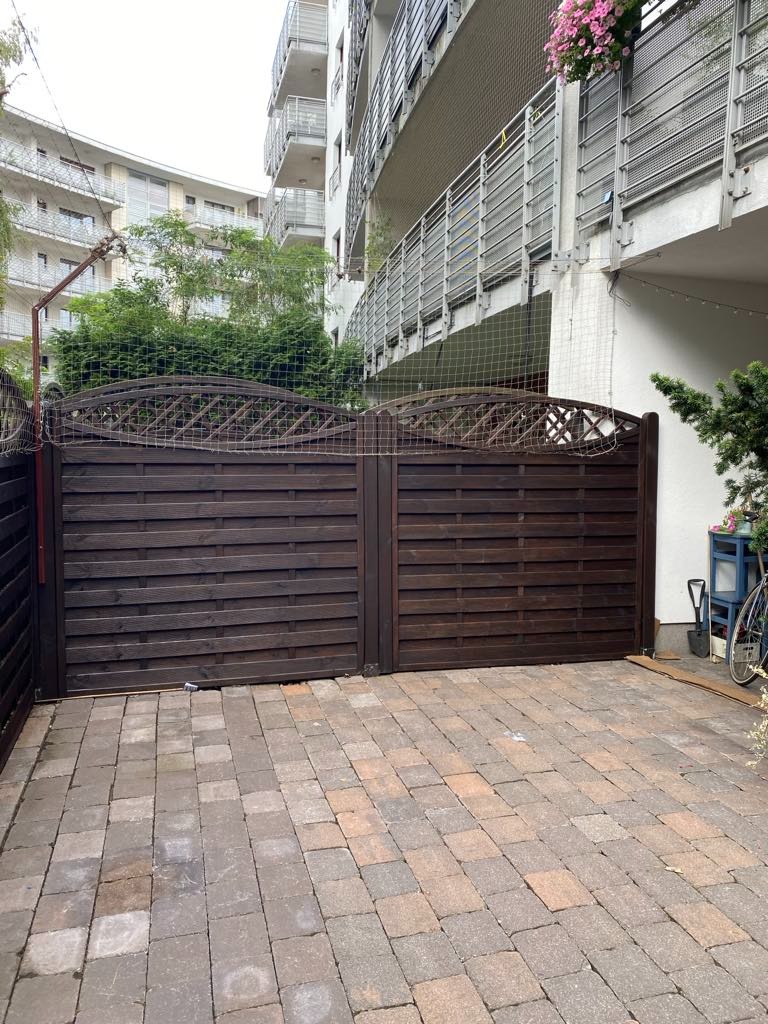 Ciemnobrązowa, drewniana brama wjazdowa na posesję z kostką brukową, na tle budynku wielorodzinnego z balkonami i zielenią. Widoczne ślady użytkowania.