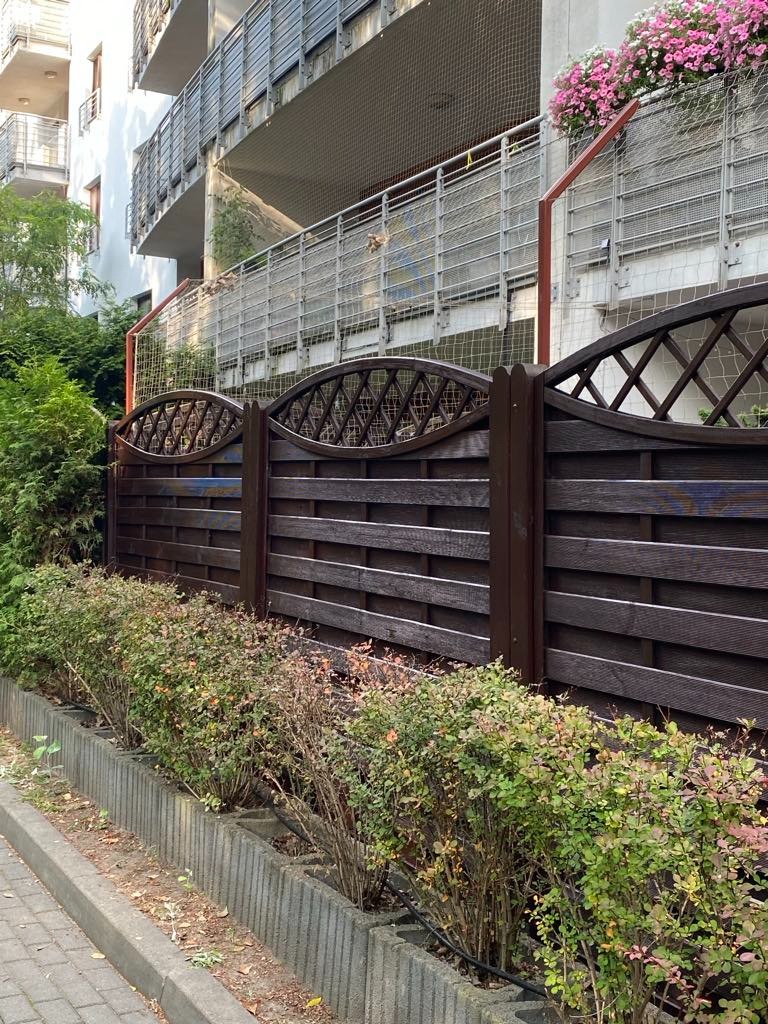 Ciemnobrązowe, drewniane ogrodzenie z ozdobnym wzorem na tle balkonu z siatką zabezpieczającą i kwiatami. Elementy architektury miejskiej i zieleni.