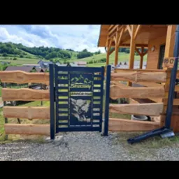 Czarna, metalowa furtka z motywem gór i jelenia, wkomponowana w drewniane ogrodzenie na tle pagórkowatego krajobrazu. Furtka z napisem 'Bieszczady'.