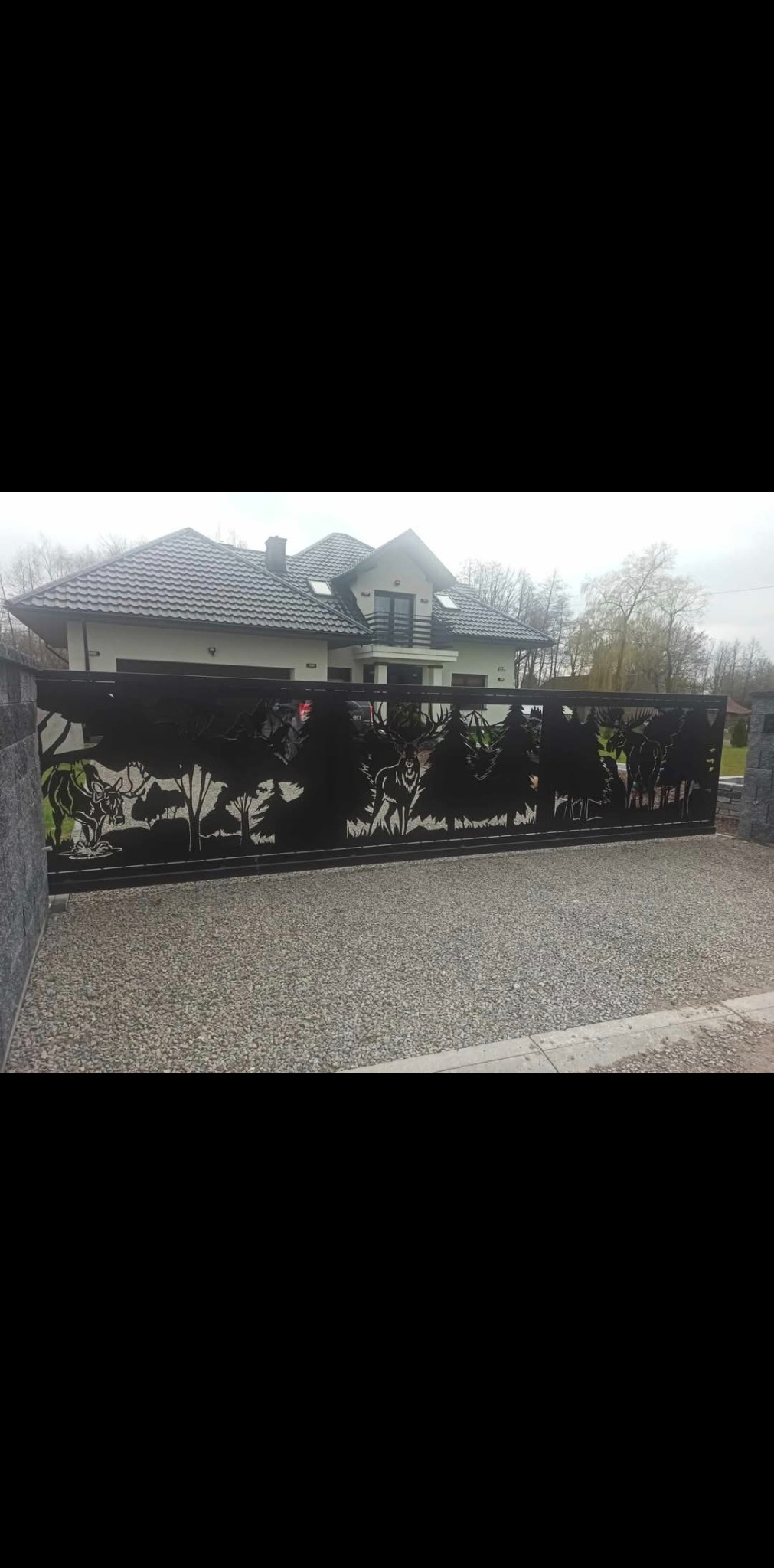 Czarna, metalowa brama przesuwna z wycinanym laserowo motywem leśnym (jelenie, drzewa) na tle jasnego domu z ciemnym dachem. Brama na żwirowym podłożu.