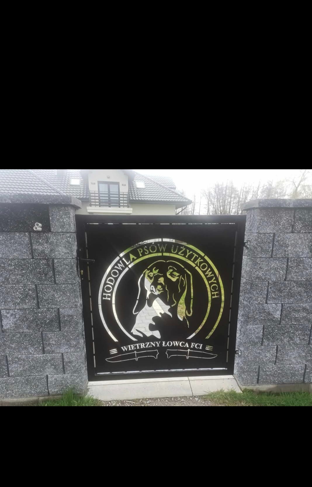 Metalowa brama wjazdowa z wyciętym laserowo logo hodowli psów użytkowych 'Wietrzny Łowca FCI'. Brama osadzona na murowanych słupkach.