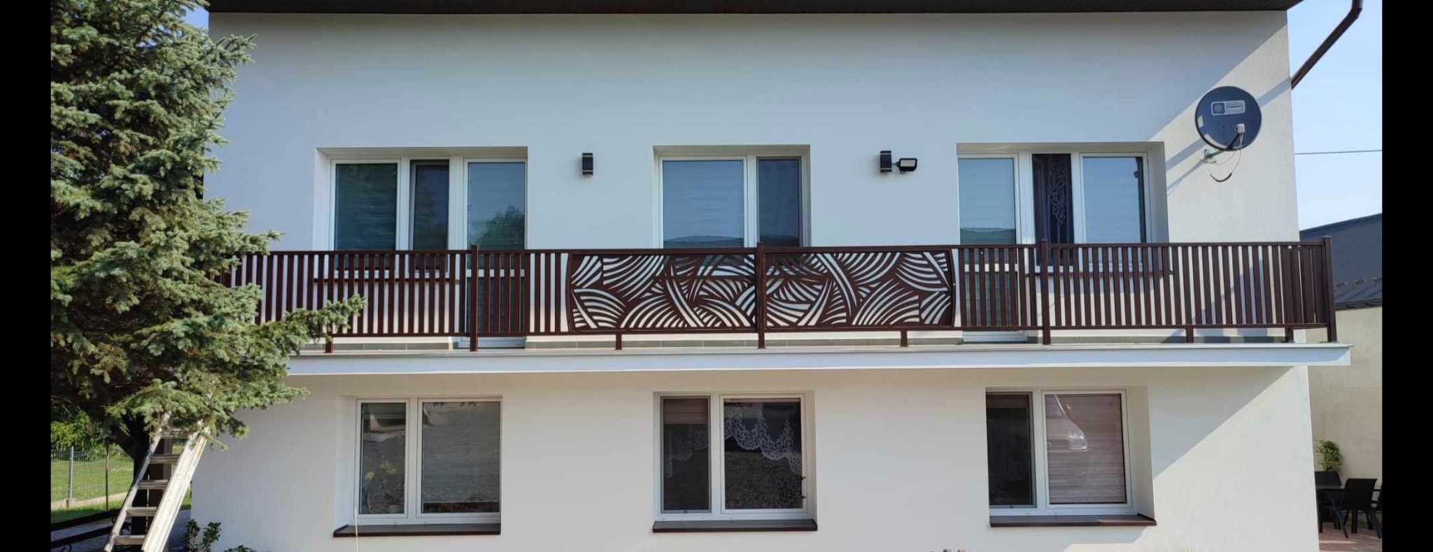 Balkon z dekoracyjną, brązową balustradą z motywem fal, widok z dołu. Elewacja budynku w Zasowie. Po lewej stronie widoczny fragment drzewa.